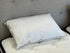 A white pillow with a thin dark piping trim, resting on a white quilted mattress against a gray tufted headboard.